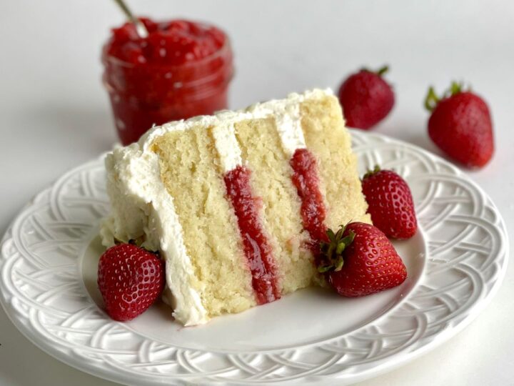A slice of white cake with strawberry filling and white frosting on a plate, garnished with fresh strawberries; a jar of strawberry preserve is in the background.