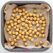 A square baking tray lined with parchment paper holds a single layer of chickpeas ready for roasting.