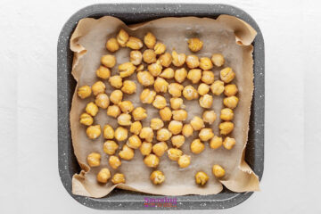 A square baking tray lined with parchment paper holds a single layer of chickpeas ready for roasting.