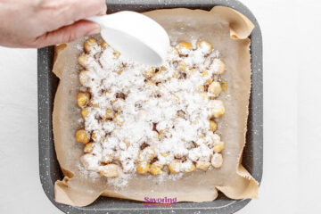 A hand sprinkles ranch dressing powder over a square baking dish lined with parchment paper and filled with chickpeas.