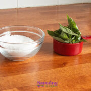 A glass bowl of coarse salt and a red measuring cup filled with fresh basil leaves on a wooden countertop.