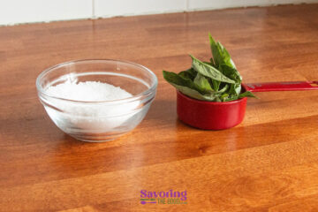 A glass bowl of coarse salt and a red measuring cup filled with fresh basil leaves on a wooden countertop.