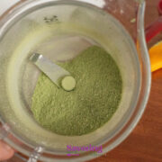 A blender cup containing Basil Salt, viewed from above, with a metal blade in the center and kitchen utensils in the background.