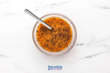 A glass bowl filled with an orange-brown liquid, topped with ground black pepper, sits on a white marble surface. A metal spoon rests in the bowl.