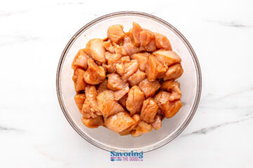 A glass bowl filled with raw, marinated chicken pieces on a white surface.