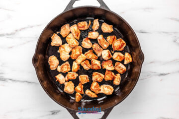 Bite-sized pieces of cooked chicken are shown in a cast iron skillet on a white marble surface.