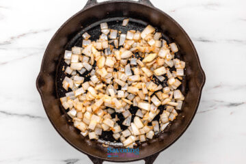 Diced onions sautéing in a cast iron skillet on a white marble countertop.
