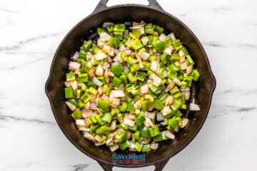 Diced green bell peppers and onions are sautéing in a black cast iron skillet on a white marble countertop.