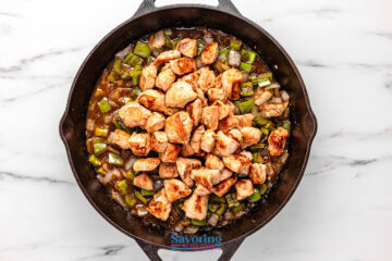 A cast iron skillet filled with cooked diced chicken, chopped celery, and onion in a brown sauce, seen from above on a marble surface.