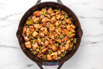 Overhead view of cooked diced chicken with green bell peppers and onions in a black skillet on a white marble surface.