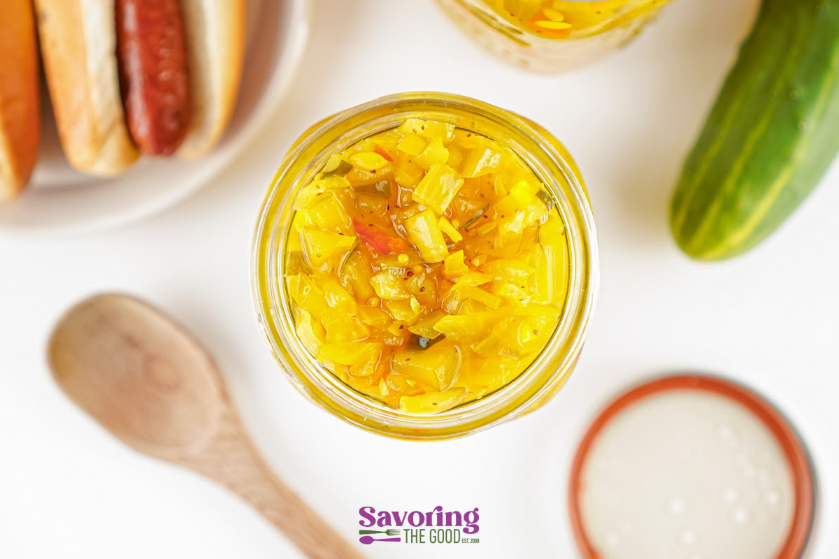 An open jar of yellow relish sits on a white surface near a hot dog, cucumber, wooden spoon, and a lid.
