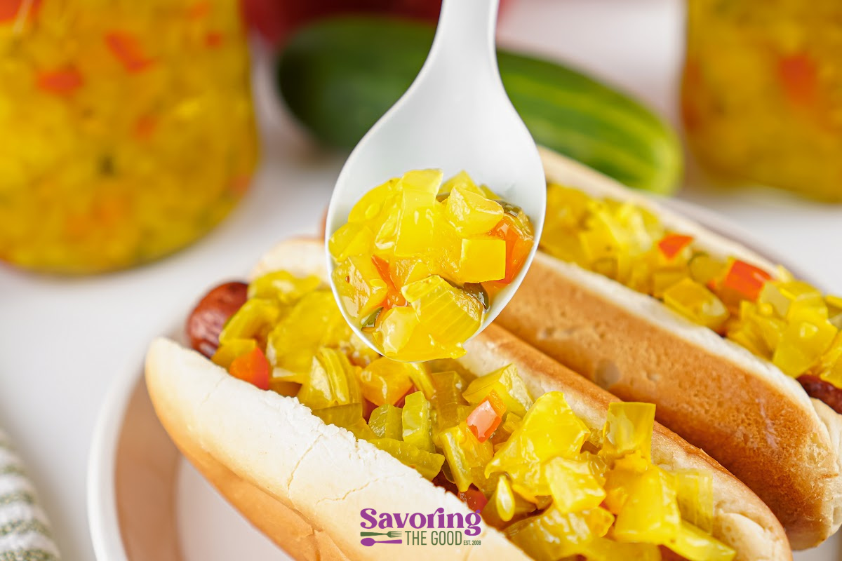 A spoonful of yellow relish is being added to two hot dogs in buns, with relish jars and a pickle in the background.