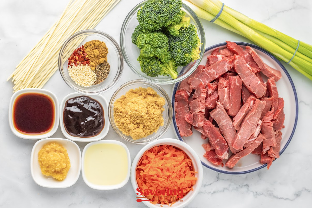 Raw ingredients for a stir fry are arranged on a white surface, including sliced beef, broccoli, noodles, carrots, sauces, spices, ginger, and green onions for Beef lo mein.