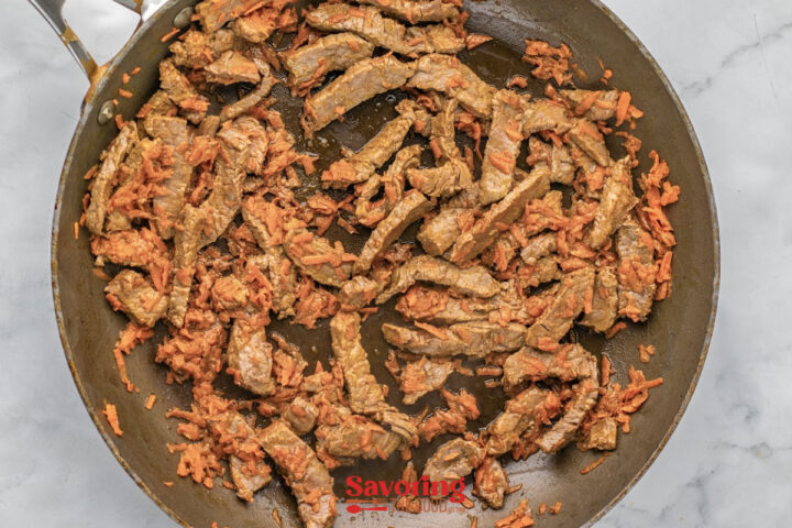 Beef strips and grated carrots cooking in a skillet, viewed from above on a white surface.