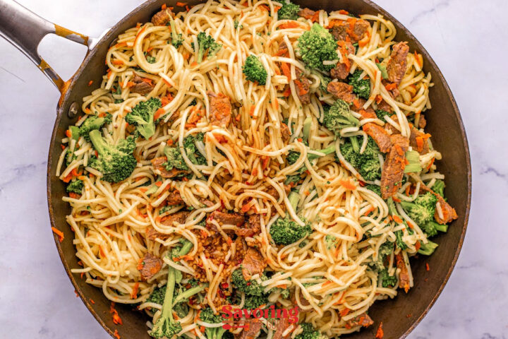 A skillet filled with cooked spaghetti noodles, broccoli florets, sliced beef, and shredded carrots, all mixed together.