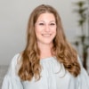 A woman with long brown hair wearing a light-colored blouse smiles at the camera in a softly lit indoor setting, offering a welcoming glimpse into the post author's personality.Sarah Mock headshot 100by100.