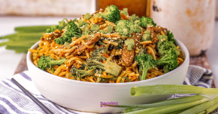 A bowl of stir-fried noodles with beef, broccoli, and chopped green onions, garnished with sesame seeds, on a striped cloth with green onions beside the bowl.