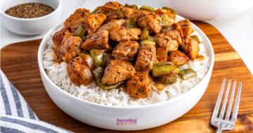 A bowl of white rice topped with cooked, seasoned chicken pieces, diced green bell peppers, and onions, placed on a wooden board with a fork and a bowl of pepper nearby.