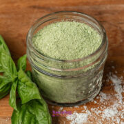 A small glass jar filled with Basil Salt sits on a wooden surface next to fresh basil leaves and a sprinkle of coarse salt.