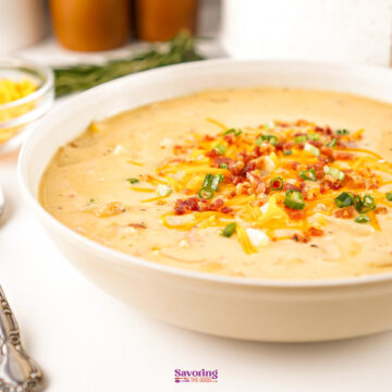 A bowl of cheesy potato soup topped with shredded cheese, bacon bits, and chopped green onions sits on a white surface.
