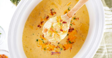 A wooden spoon holds a serving of Cheesy Potato soup garnished with chopped green onions and crispy bacon bits in a white bowl.