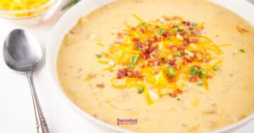 A bowl of creamy cheesy potato soup topped with shredded cheese, crumbled bacon, and chopped green onions; a spoon is placed beside the bowl.