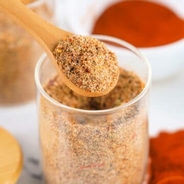A wooden spoon holds a portion of seasoned dry spice mix above a glass jar filled with the same jerk seasoning recipe; a bowl of red spice is in the background.