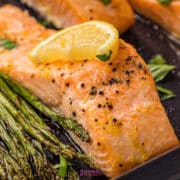 A sheet pan salmon fillet cooked to perfection, topped with a lemon wedge and served alongside roasted asparagus on a dark tray.
