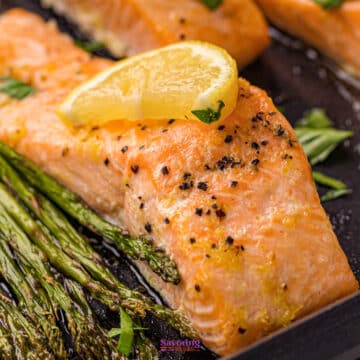 A sheet pan salmon fillet cooked to perfection, topped with a lemon wedge and served alongside roasted asparagus on a dark tray.