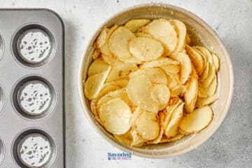 A bowl of thinly sliced potatoes seasoned with grated cheese sits next to an empty muffin tin on a gray countertop.