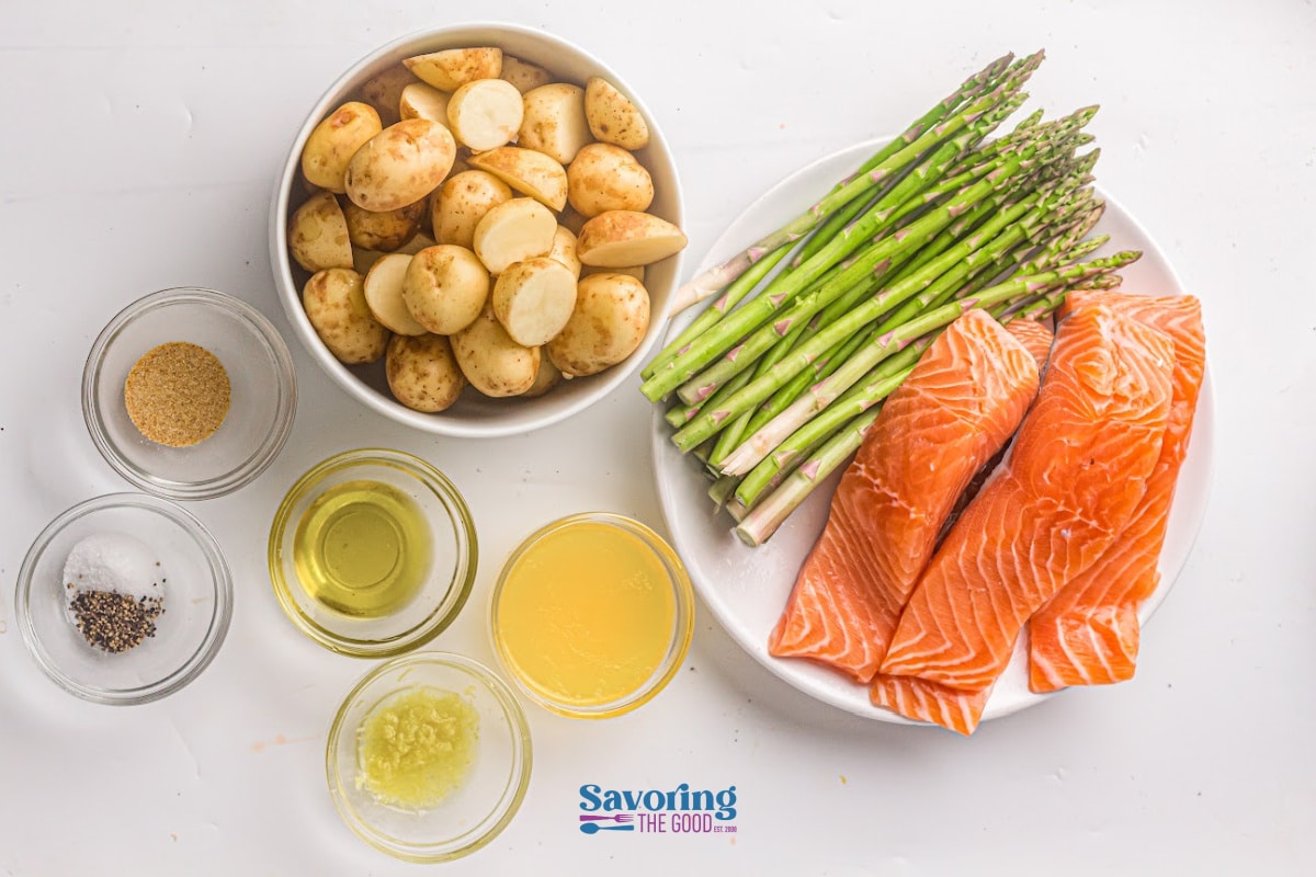 Sheet pan salmon with raw fillets, asparagus, halved baby potatoes, and small bowls of seasonings, oil, lemon juice, and minced garlic all arranged on a white surface.