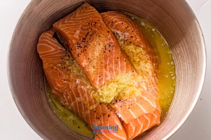 Two raw salmon fillets in a metal bowl with melted butter, minced garlic, black pepper, and seasoning—perfect for preparing flavorful sheet pan salmon.
