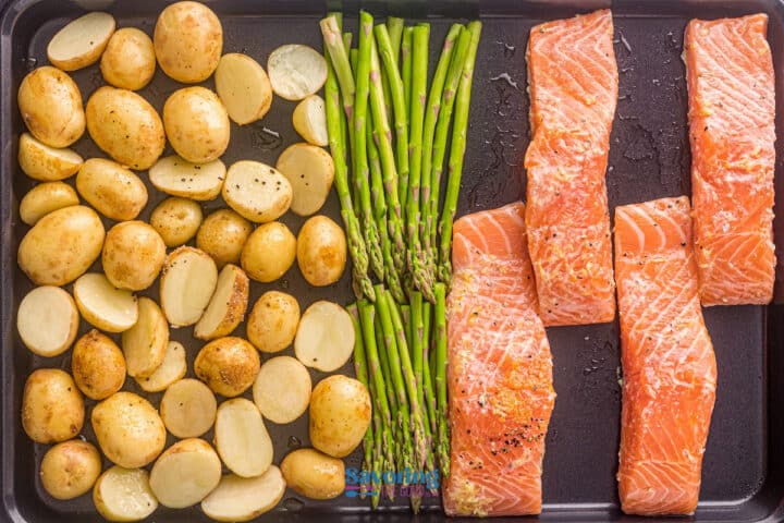 A sheet pan salmon dinner featuring seasoned fillets, halved baby potatoes, and asparagus spears arranged in separate sections, ready for cooking.