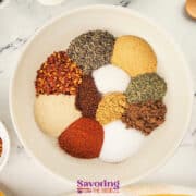 A white bowl contains various jerk seasoning ingredients arranged in separate piles-black pepper, red pepper flakes, paprika, salt, and dried herbs-on a marble surface with other bowls nearby.