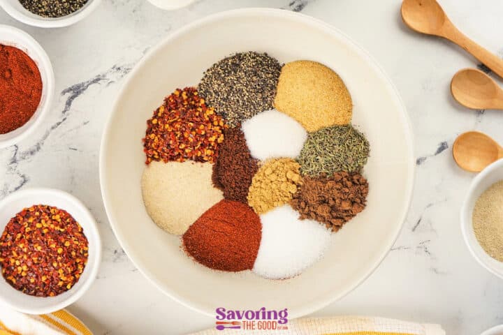 A white bowl contains various jerk seasoning ingredients arranged in separate piles-black pepper, red pepper flakes, paprika, salt, and dried herbs-on a marble surface with other bowls nearby.