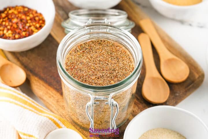 A glass jar filled with a homemade jerk seasoning recipe sits on a wooden board, surrounded by wooden spoons and bowls containing other spices.