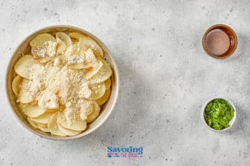 A bowl of sliced potatoes topped with grated cheese next to small bowls of herbs and a brown liquid on a light gray surface.