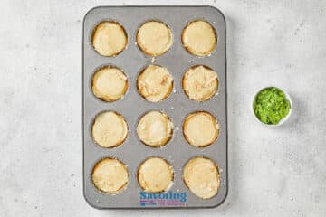 A muffin tin with twelve unbaked dough rounds inside, next to a small bowl of chopped green herbs on a light surface.