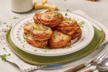 A white plate holds a stack of golden, crispy potato stacks garnished with chopped herbs, set on a green placemat beside a fork and knife.