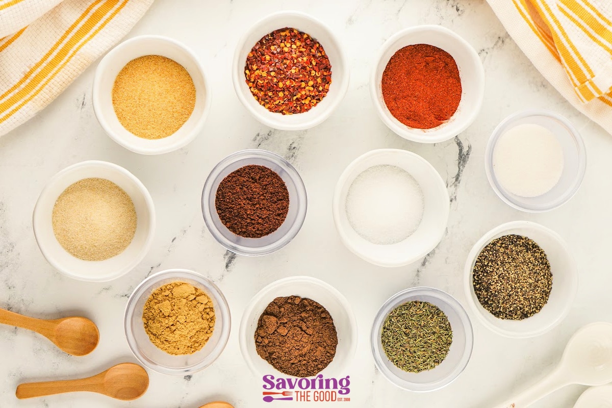 Twelve small bowls containing various ground spices and herbs are arranged on a marble surface with two wooden spoons nearby.