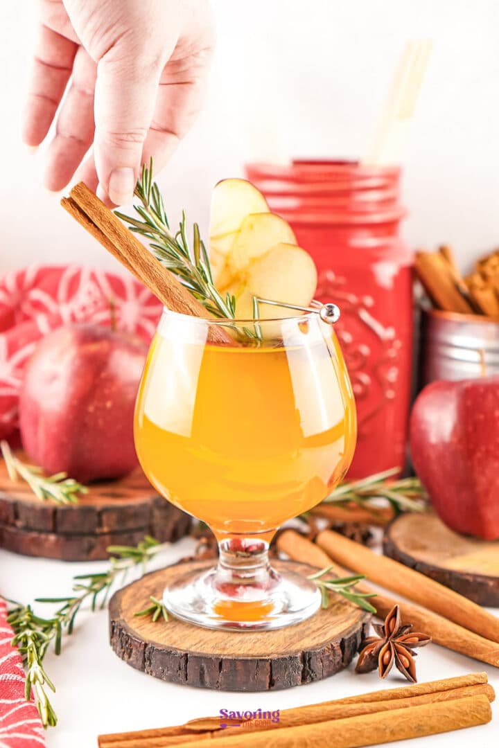 A hand garnishes a glass of orange-colored beverage with a cinnamon stick and rosemary sprig; apples, cinnamon sticks, and a red jar are in the background.