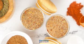 Two glass jars filled with mixed dry spices, including a homemade jerk seasoning recipe, sit on a white surface, surrounded by additional bowls and loose spices.