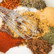 A metal whisk rests in a bowl containing assorted spices and seasonings, including vibrant piles ready for a jerk seasoning recipe.