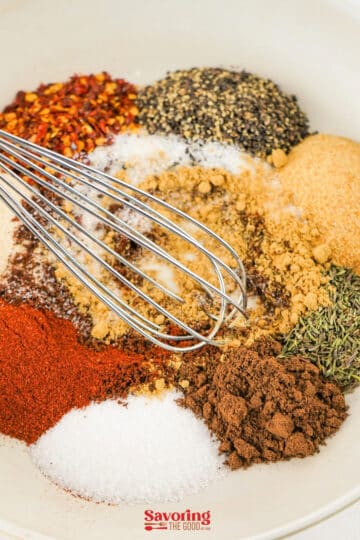 A metal whisk rests in a bowl containing assorted spices and seasonings, including vibrant piles ready for a jerk seasoning recipe.