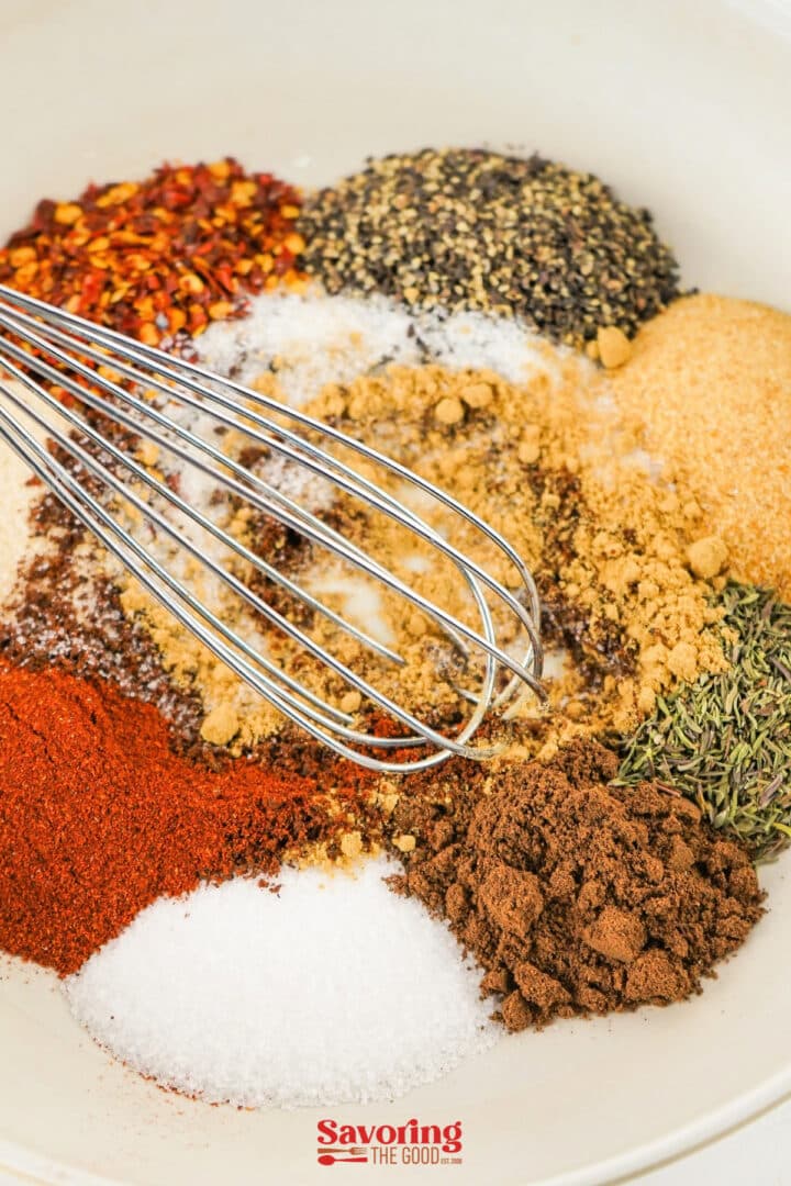 A metal whisk rests in a bowl containing assorted spices and seasonings, including vibrant piles ready for a jerk seasoning recipe.