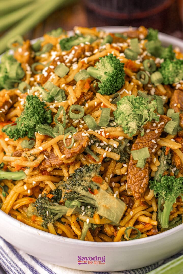 A bowl of stir-fried noodles with broccoli, green onions, sesame seeds, and slices of meat, served in a white dish.