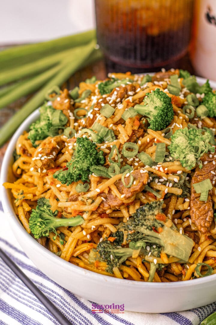 A bowl of stir-fried noodles with broccoli, sliced meat, chopped green onions, and sesame seeds, served on a striped cloth with green onions and a drink in the background.