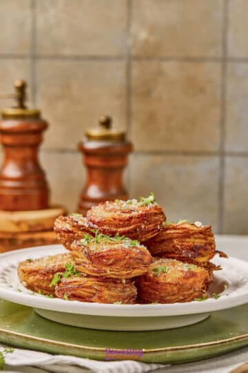 A plate of stacked crispy potato rounds garnished with herbs and coarse salt, with two wooden pepper mills in the background.