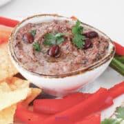 A bowl of black bean dip garnished with cilantro and beans, surrounded by tortilla chips and red bell pepper slices.