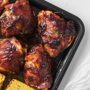 Baked chicken thighs coated in a glossy barbecue sauce, garnished with herbs, served in a black tray alongside pieces of corn on the cob.